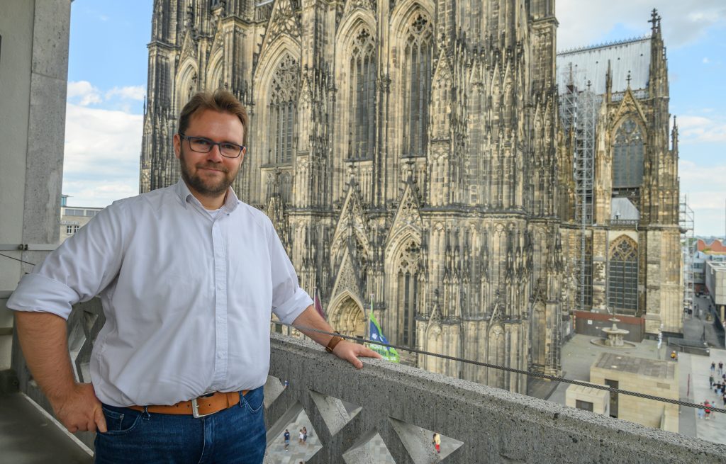 Renato Schlegelmilch, von dem der Text handelt, steht auf einem Balkon, im Hintergrund ist die Fassade des Kölner Doms zu sehen. 