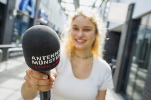 Eine junge Reporterin hält ein Mikro mit der Aufschrift "Antenne Münster" vor die Kamera.