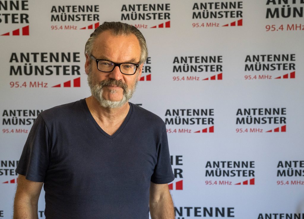 Antenne-Münster-Chefredakteur Stefan Nottmeier steht vor einer Wand mit dem Logo des Senders. 