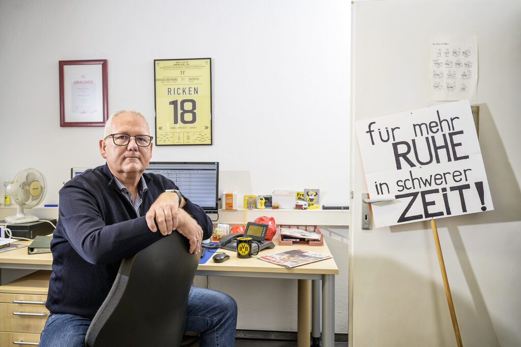 Ein Mann sitzt an einem Schreibtisch, neben ihm ein Schild "Für mehr ruhe in schwerer Zeit".