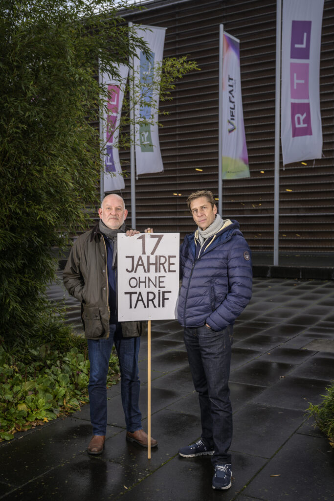 Zwei Betriebsräte von RTL News stehen in Winterjacken vor RTL Fahnen und halten ein Schild: "17 Jahre ohne Tarif".