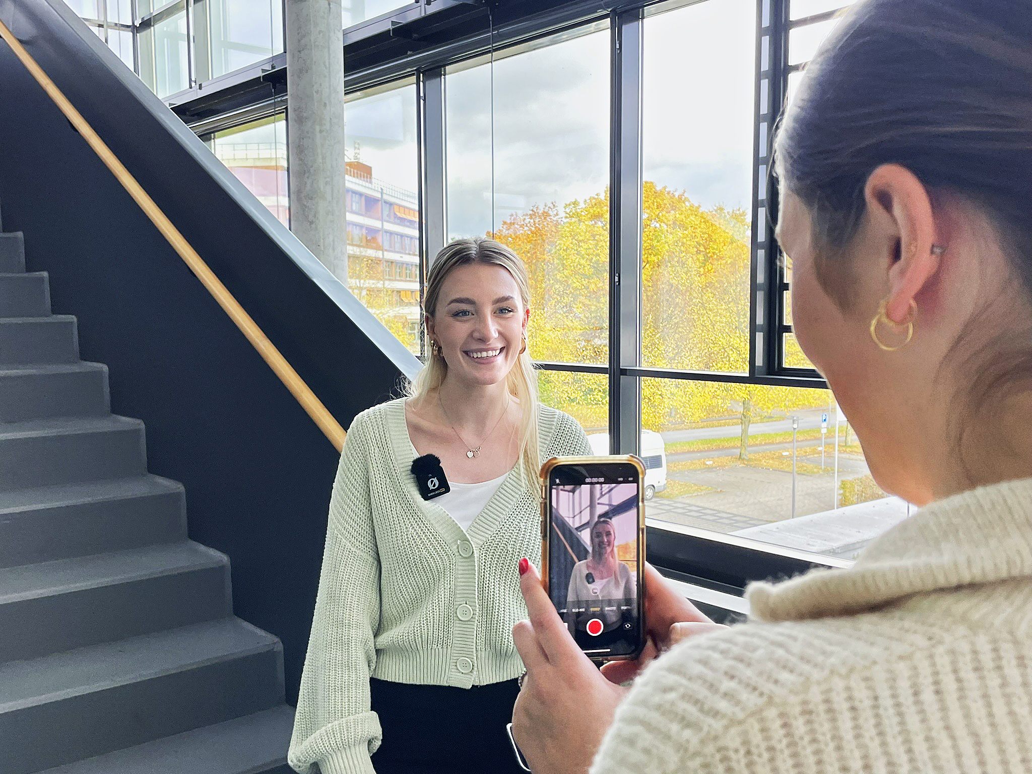 Eine Studentin filmt mit dem Handy ihre Kommilitonin, die vor einer Treppe steht.