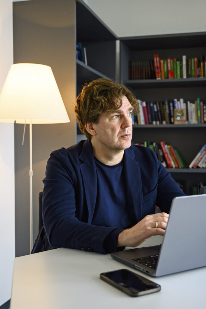 Ein Mann sitzt in seinem Arbeitszimmer am Laptop, im Hintergrund eine Bücherwand. 