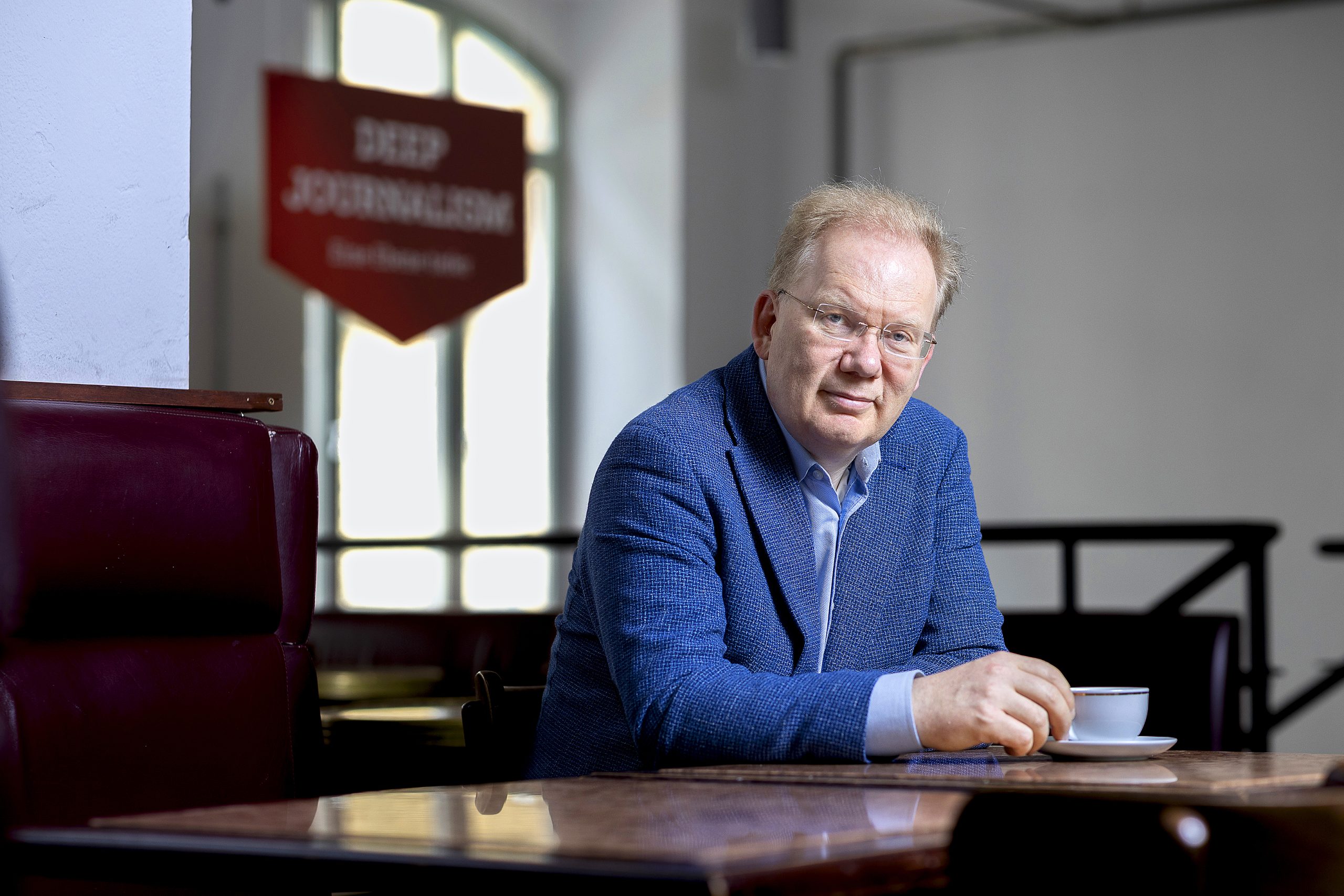 Ein Mann im Blauen Jackett sitzt mit einer Tasse Kaffee an einem Tisch und guckt in die Kamera. Hinter ihm ist unscharf ein Schild mit dem Schriftzug Deep Journalism zu erennen.