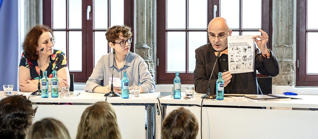 Ein Podium mit zwei Frauen und einem Mann, der ein Dokument hochhält.