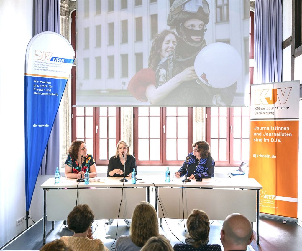 Über die Köpfe des Publikums hinweg ist ein Panel mit drei Frauen zu sehen. Über ihnen wird auf einer Leinwand ein Bild von den Protesten in Belarus projiziert.