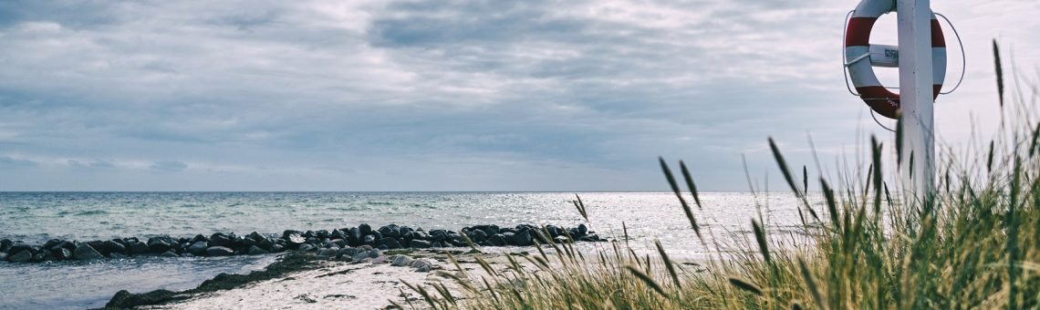 Ein Strand unter wolkigem Himmel, rechts steht ein Pfhal mit einem Rettungsring