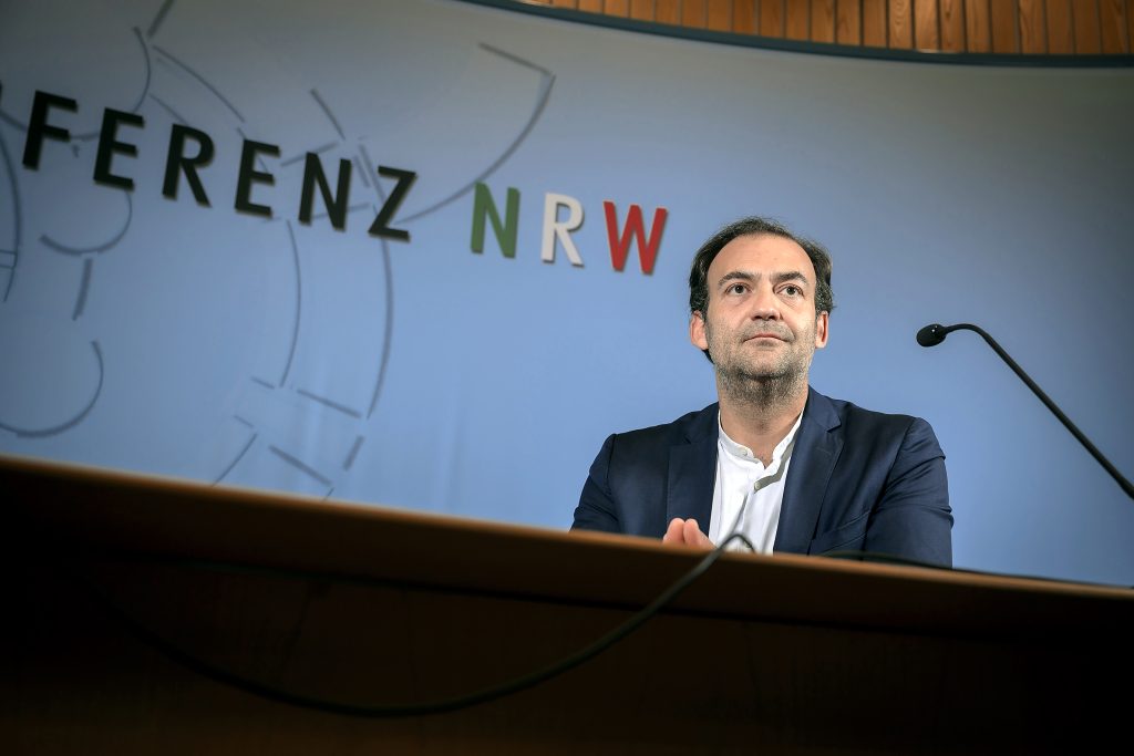 Ein Mann sitzt an einem Pult, er ist von schräg unten fotografiert. Hinter ihm ist ein Teil des Namens Landespressekonferenz NRW zu erkennen. 