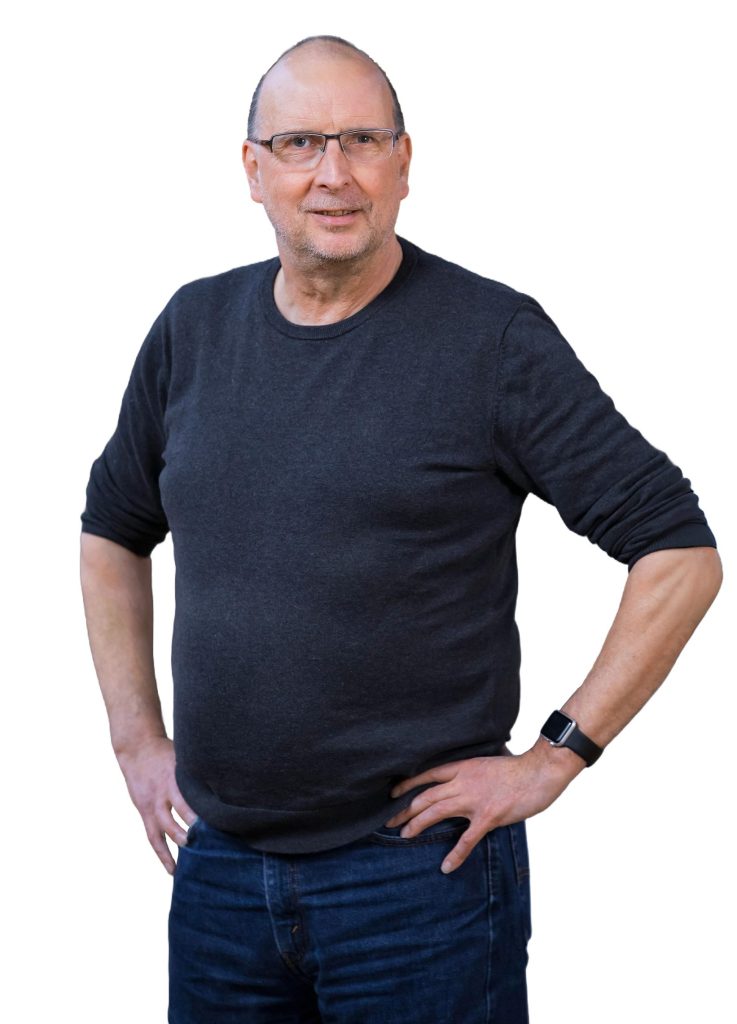 Ein Mann mit sehr kurzen Haaren und Brille, der die Arme in die Hüften stützt und freundlich lächelt. Er trägt Jeans und einen Pulli mit hochgekrempelnten Ärmel.