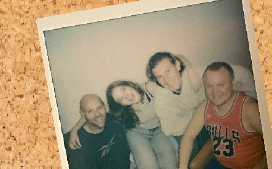 Auf einer Pinwand aus Kork hängt ein Polaroid in verblassten Farben. Zu sehen sind drie jungen Männer und eine junge Frau, die eing beieinander hocken.