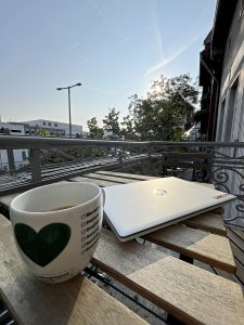 Ein Balkontisch mit Kaffeebecher und zugeklapptem Laptop. Der Blick geht auf etwas städtisches Grün und den blauen Himmel.