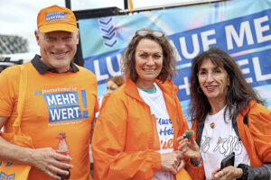 Ein Mann in einem organgen T-Shirt und zwei Frauen in orangen Regenjacken lächeln in die Kamera.