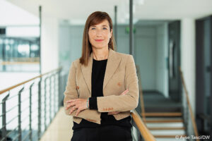Ein Frau mit langen dunklen Haaren steht mit verschränkten Armen in einem hellen Gang. Links von ihr ist ein Geänder zu erkennen.