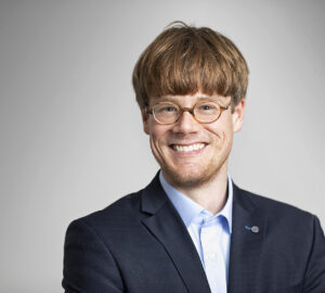 Ein junger Mann mit Brille und Bart lächelt in die Kamera. Er trägt ein helles Hemd mit offenenm Kragen nd darüber ein Jacket.
