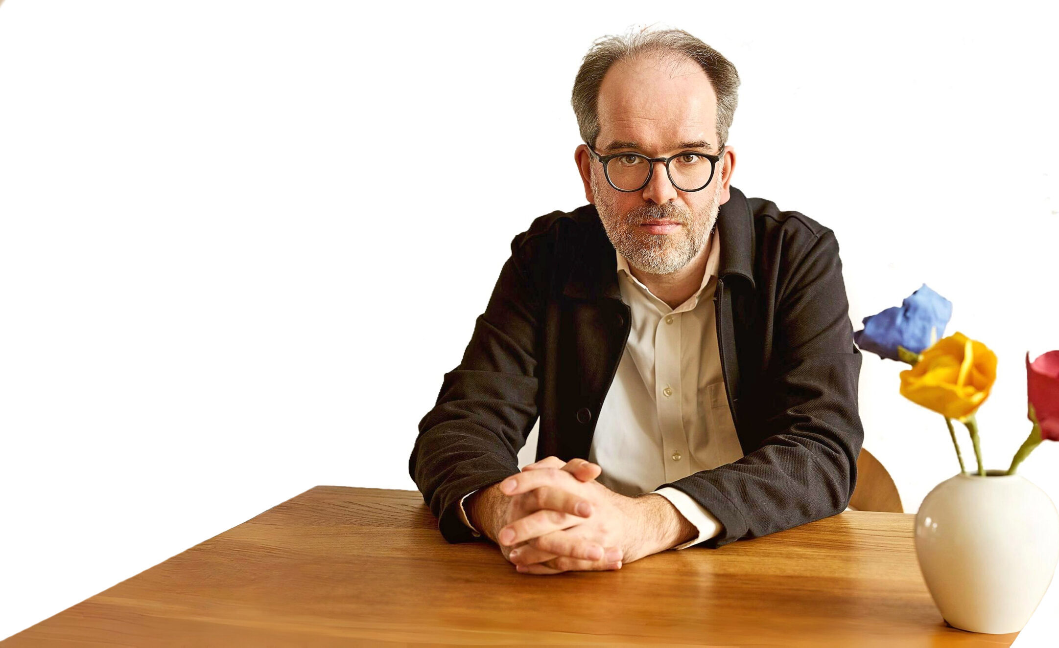 Ein Mann mit Brille und skeptischem Gesichtsausdruck sitzt an einem Tisch. Er trägt ein helles Hemd und ein dunkles Jackett. Neben ihm steht eine Vase mit Papierblumen.