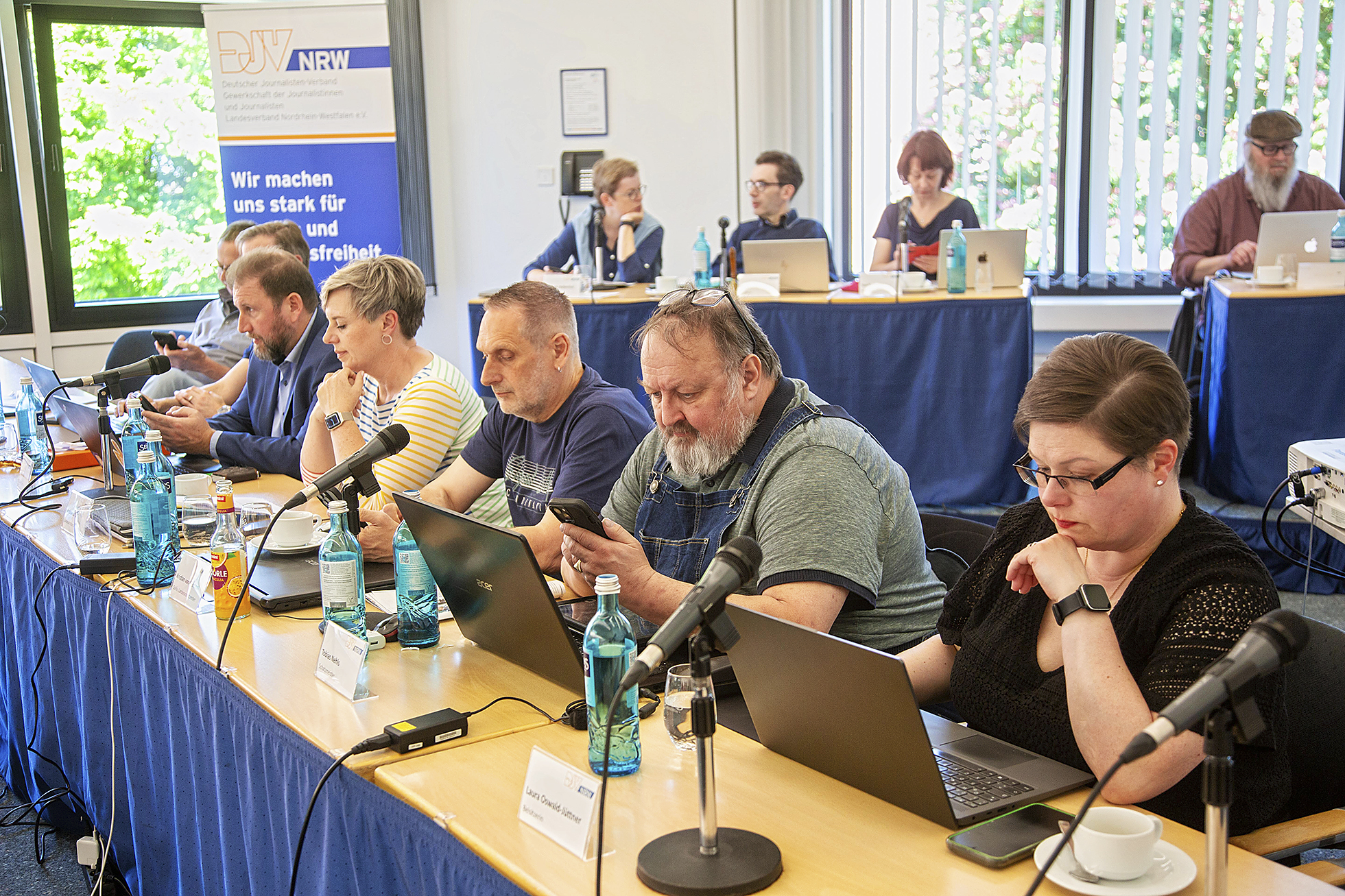 An einem langen Tisch sitzen insgesamt sieben Frauen und Männer, die konzentriert auf Laptops und Tablets schauen. Dahinter zwei Tische mit insgesamt vier Personen.