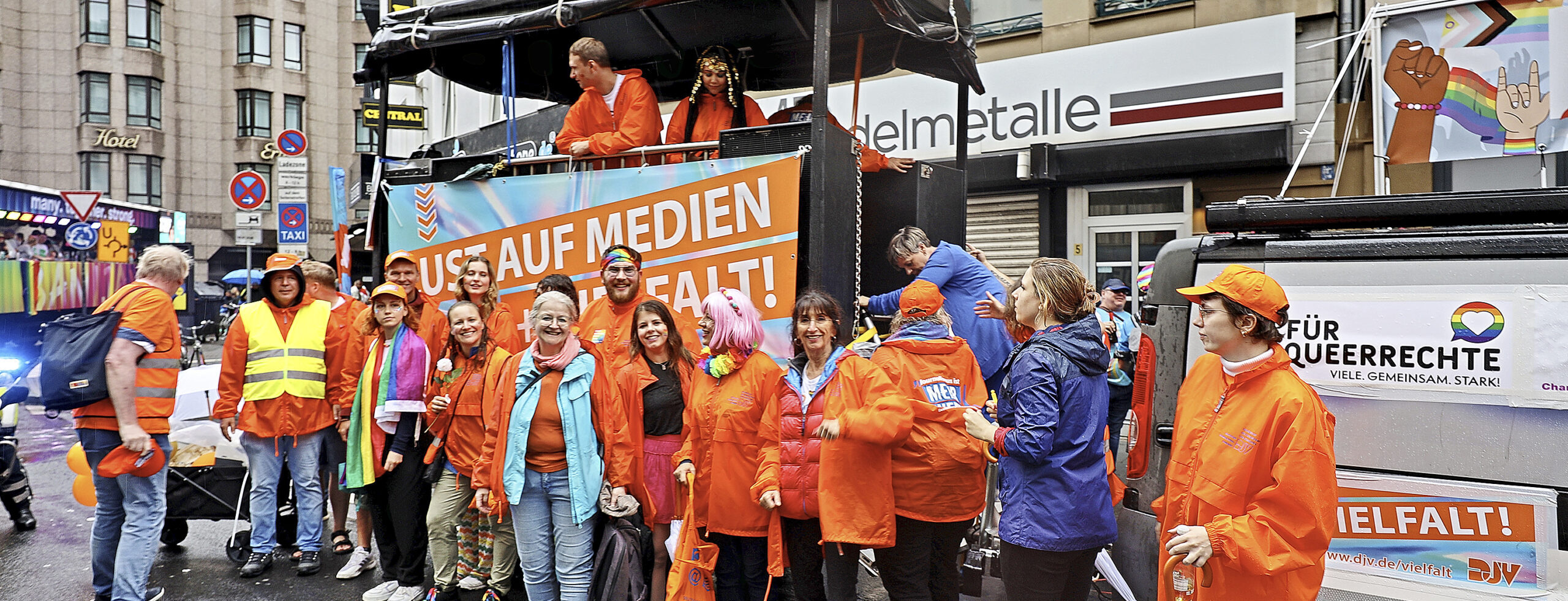 Ein Gruppe von Menschen steht vor einem Wagen mit Banner. Auf dem Wgen stehen zwei weitere Personen. Die meistenr in der Gruppe tragen orange Regenjacken.