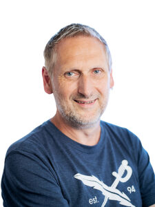 Ein Mann in einem blauen T-Shirt lächelt in die Kamera.