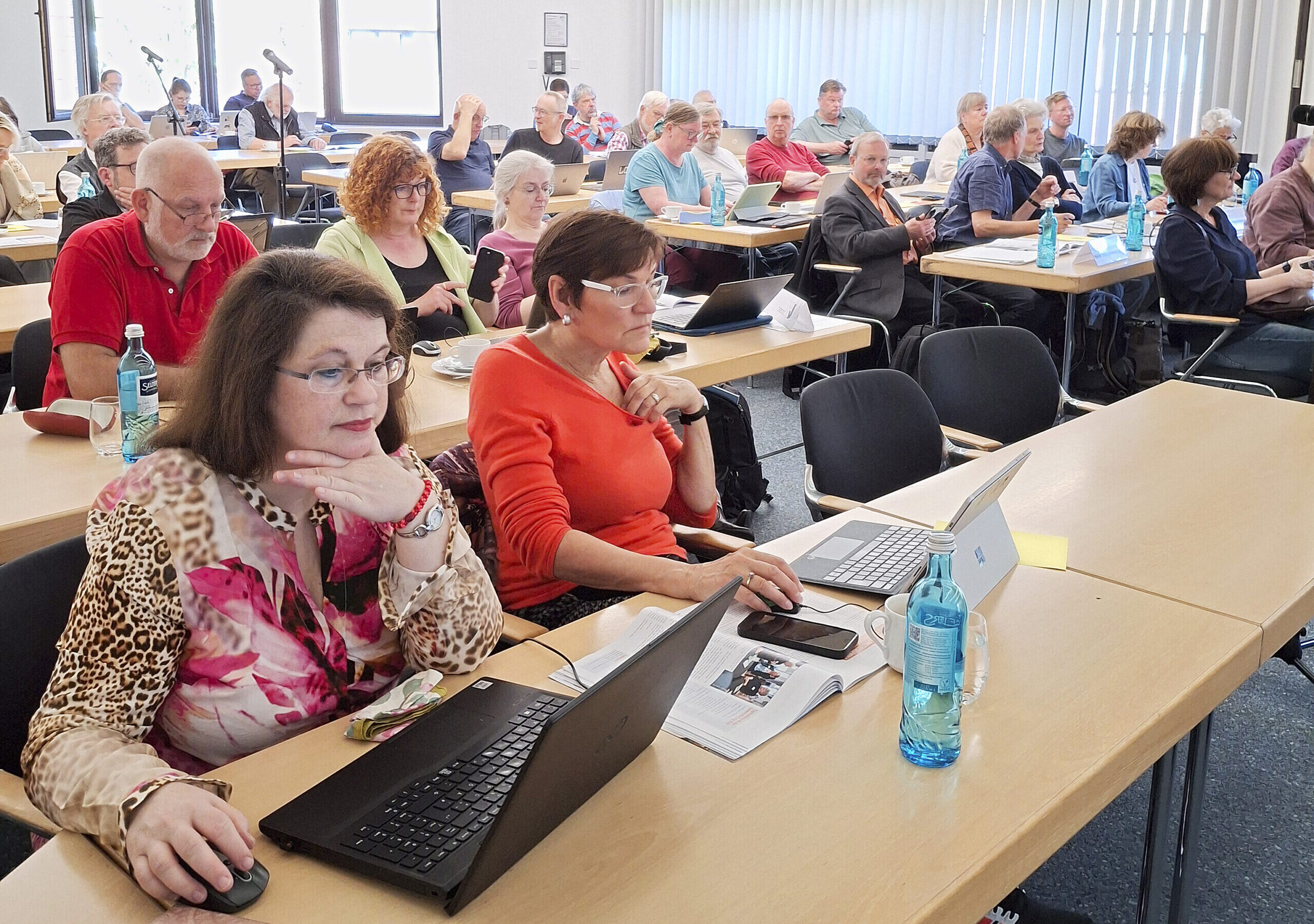 Blick in einen Tagungssaal. Die Menschen sitzen in Reihen an langen Tischen. Einige chauen in Laptops.