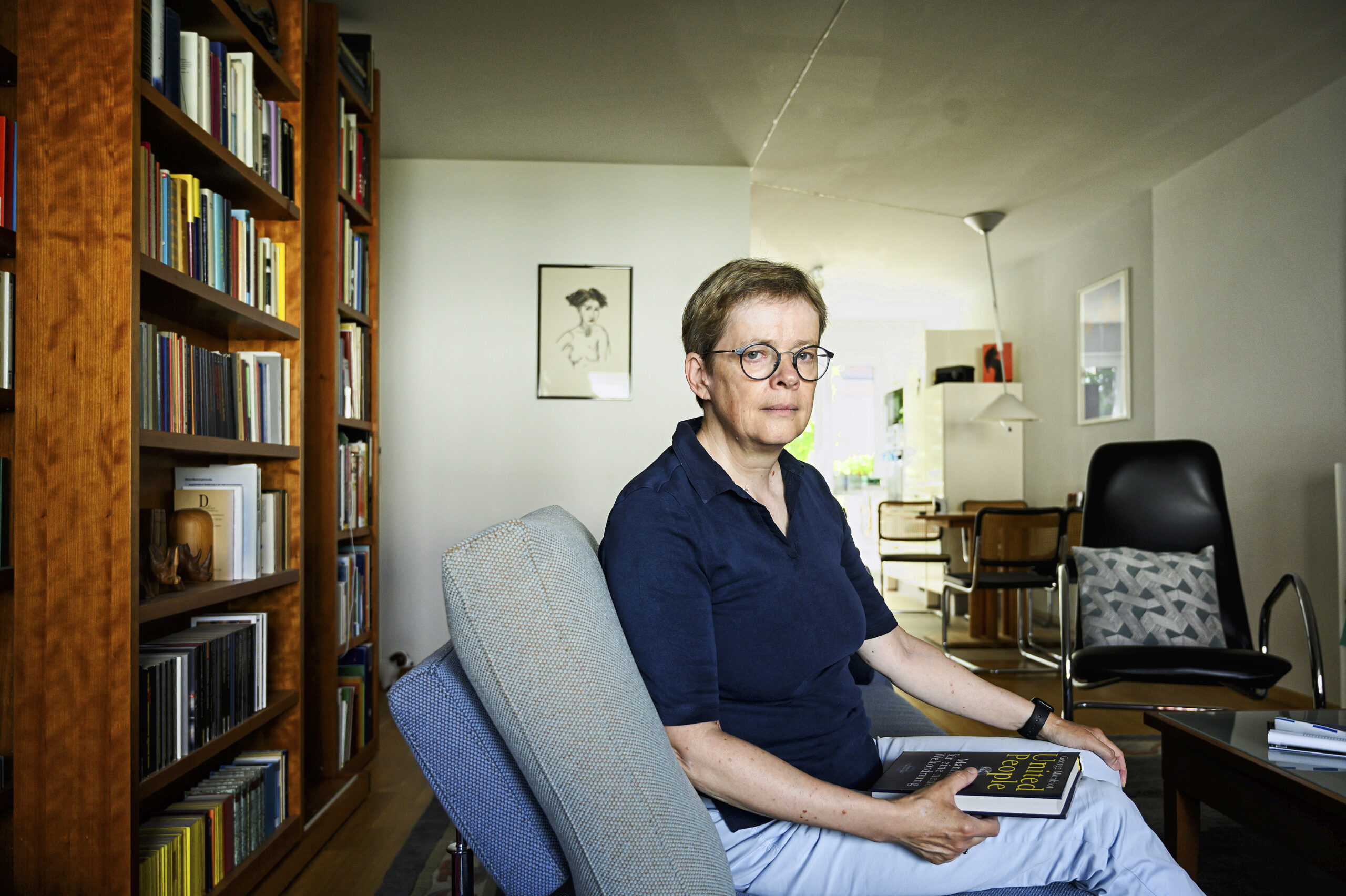 Eine Frau schaut seitlich mit ernstem Gesicht in die Kamera. Sie ist Brillenträgerin und hat kurze mittelbrauche Haare. Die Frau sitzt auf einem Sessel und hat ein buch auf den Knien liegen, im Hintergrund ist ein rprivater Raum zu shen, links ein gut gefülltes Bücherregal.