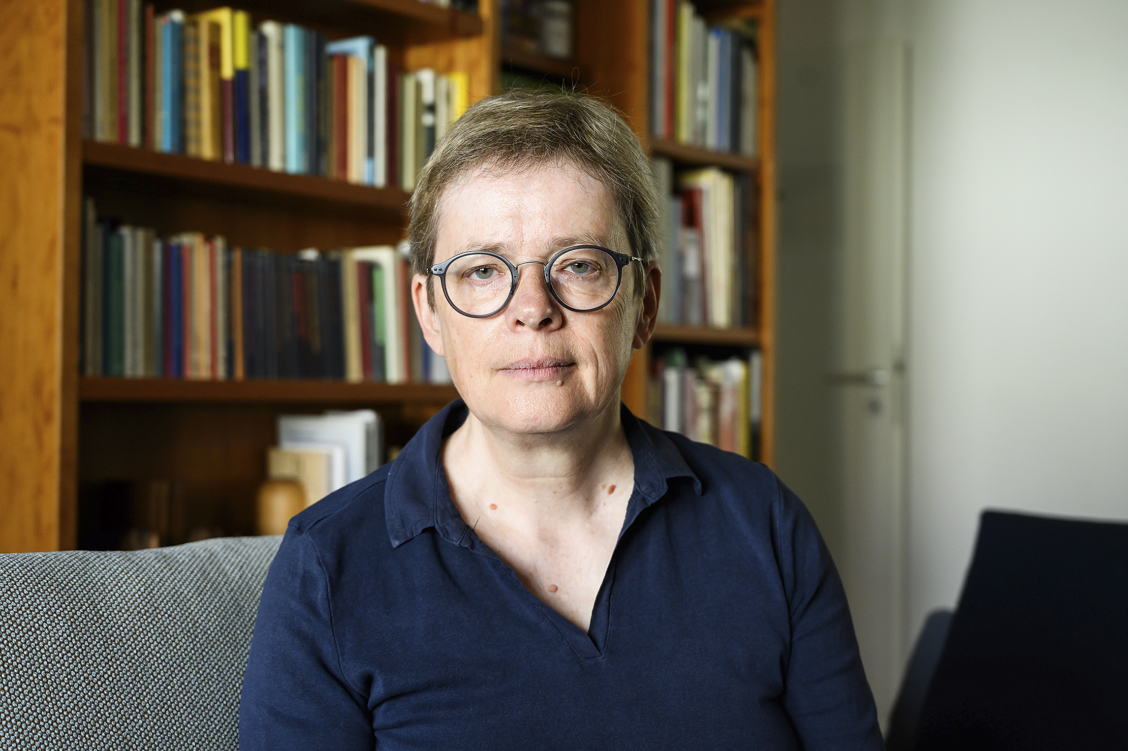 Eine Frau schaut mit ernstem Gesicht in die Kameera. Sie ist Brillenträgerin und hat kurze mittelbrauche Haare. Sie sietzt auf einem Sessel, im Hintergrund ist ein Bücherregal zu erkennen.