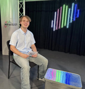 Ein Student in heller Kleidung sitzt in einem Studio vor schwarzem Hintergrund. Vor seinen Knien steht ein Kasten mit iner Oberfläche, die die verschiedenen Farben leuchtet. Rechts hinten an der Wand ist eine Lecuchtgrafik mit dem Umriss von NRW, sie besteht aus Leuchtstäben in mehreren Farben. Links hinter ihm ist eine helle Säule mit dem Schriftzug NRWision.