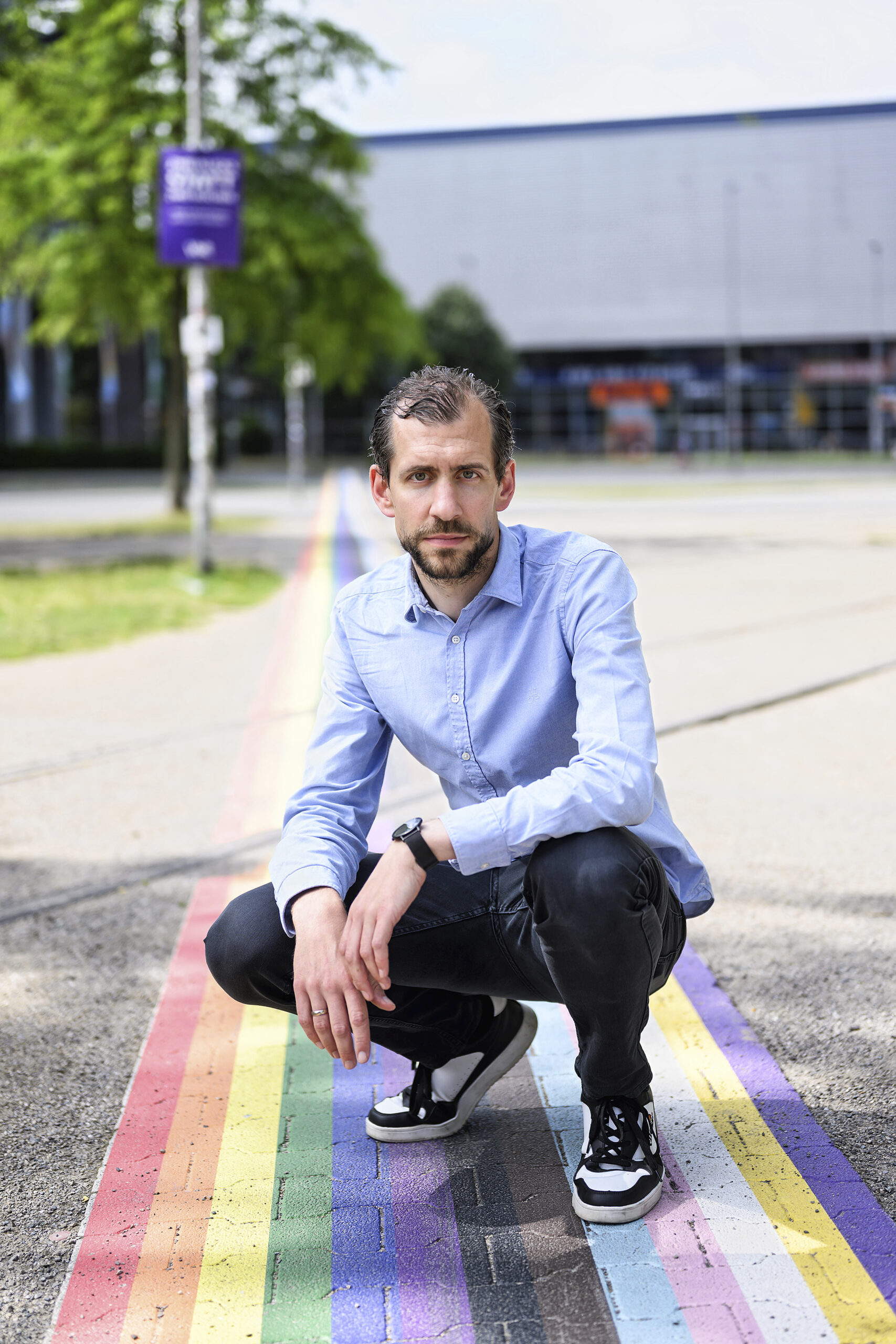 Ein dunkelhaariger Mann mit Bart hockt auf einem Radweg, der in Regenfogenfarben gestrichen ist. Er trägt ein hellblaues Hemd, eine dunkle Hose und Sneaker.