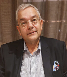 Ein Mann mit grauen Haaren und Brille schaut lächelnd in die Kamera. Er trägt ein helles Hemd mit offenen Kragen und ein dunkles Jackett mit Einstecktuch.
