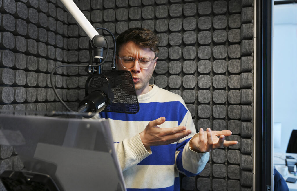 Ein junger Mann mit lockigen brauen Haaren, einer Brille und einem blau weiß gestreifeten Pulli steht in einem Aufnahmestudio. Am Gesicht und den gestikulierenden Händen sieht man, dass er sehr engagiert etwas einspricht.
