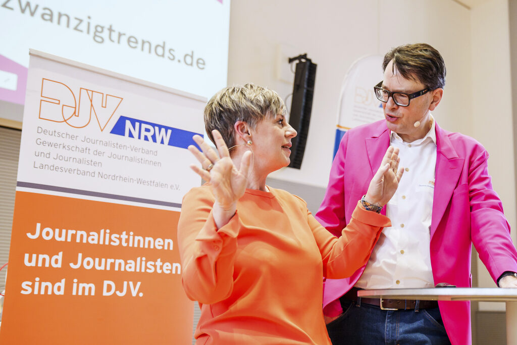Ein Frau in orangem Oberteil untrhält sich gestikulierend mit einem Mann, der ein pinkes Jacket und eine dunkle Brille trägt. Die szene ist leicht von unten fotografiert, im Hintergrund ist ein Aufsteller mit dem Logo des DJV-NRW zu erkennen.