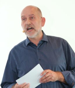 Ein Mann im blauen Hamd spricht engagiert in das Mikro eines Headsets. In den Händen hält er eine querformatiges Baltt mit Notizen.