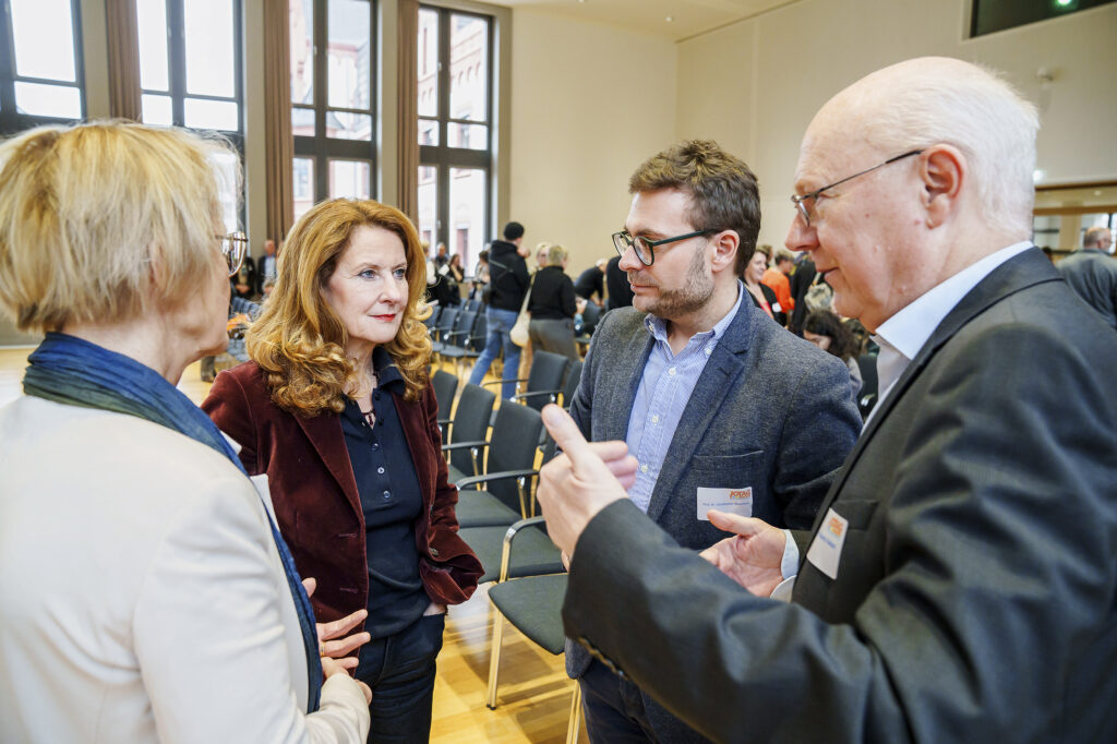 In einem großen Raum mit Stuhlreihen stehen zwei Frauen und zwi Männer zusammen und unterhalten sich.
