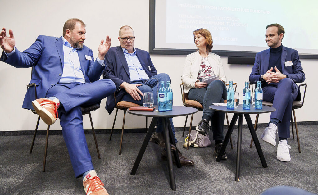 Eine Podiumsdiskussion: Links sitzt ein Mann im blauen Anzug mit überschlagenen Beinen und erklärt gestikulierend etwas. Rechts neben ihm zwei Männe und eine Frau, die zuhören.