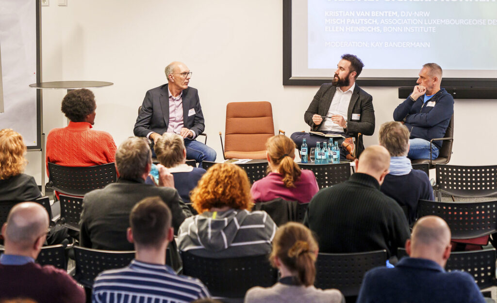 In einem größeren Seminarraum geht der Bilick üer die Hinterköpfe des Zuschauenden hiweg auf eine Podium mit drei Männern.