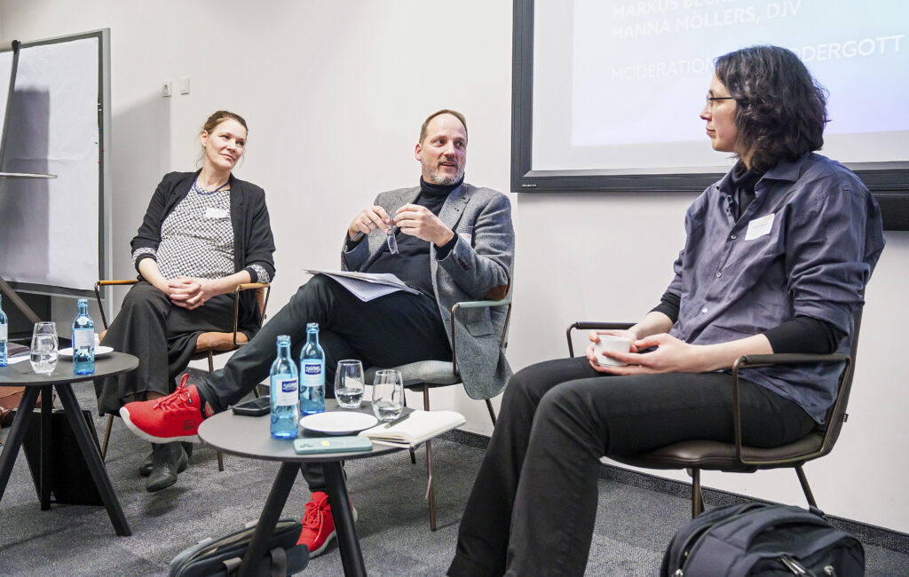 Auf einem Podium diskutieren drei Personen, links und rechts außen jeweils eine Frau, in der Mitte ein Mann.