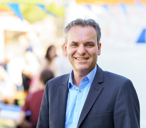 Brustbild eines lächelnden Mannes. Er hat dunkle Haare und trägt ein blaues Hemd unter grauem Jacket. Der unscharfe Hintergrund ist sommerlich hell.