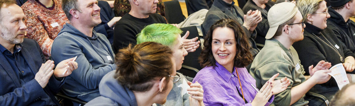 Bei einer Veranstaltung sitzen Menschen von jung bis alt eng in Zuschauerreihen. Der Blick fällt vorne links auf eine Gesprächssituation zwischen zwei jungen Frauen und einem jungen Mann, der sich zu ihnen herüberbeugt.