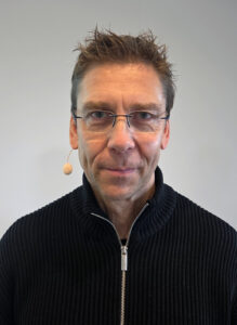 Ein Mann mit Brille und Headset schaut ernst in die Kamera. Er trägt einen dunklenPulli mit Reißverschluss.