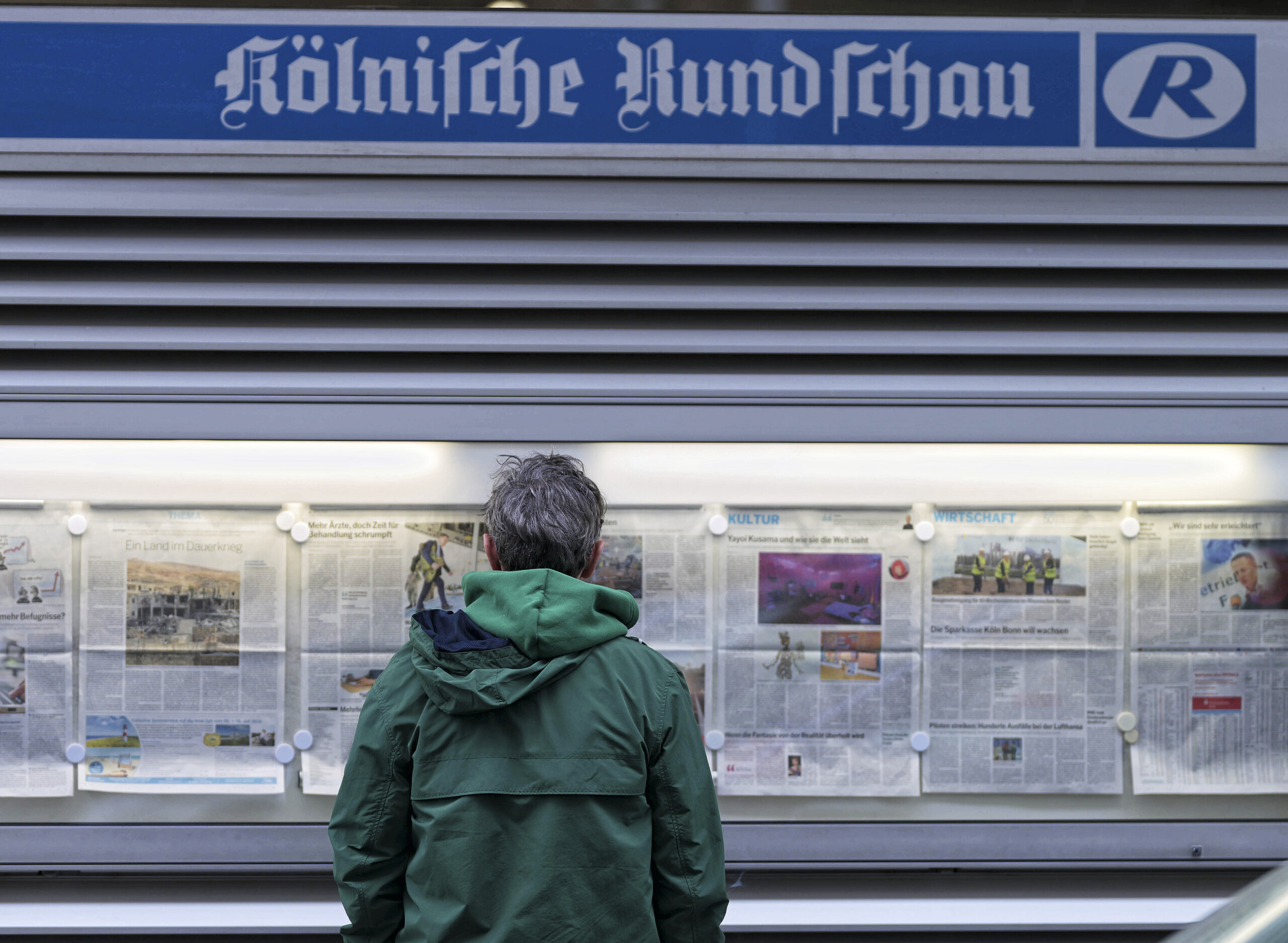 An einer Wand steht ein großer Schriftzug "Kölnisch Rundschaut. Darunter ein beleuchteter Glaskasten, in dem die Seiten der aktuellen Ausgabe der Tagesteeitung ausgestellt sind. Davor steht ein Mensch in einer grünen Jacke mit dem Rücken zu Betrachter und betrachtet die Seiten.
