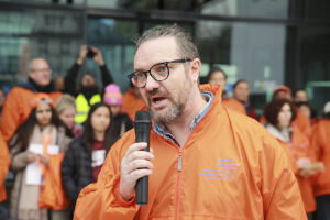 Ein Mann mit Brille und in einer orangen Jacke spricht bei einer Versammlung im Freien engagiert in ein Mikro. 