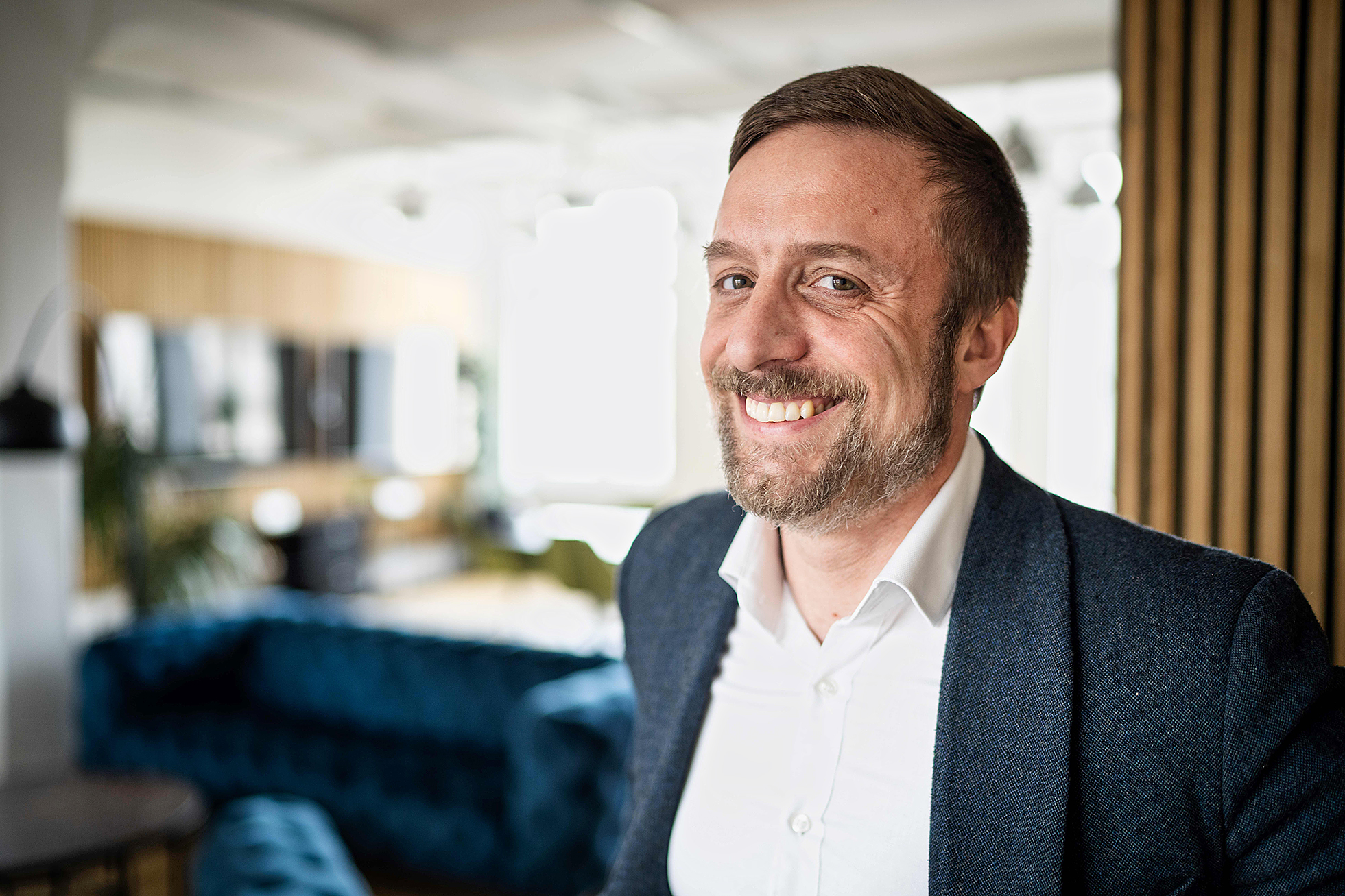 Ein Mann mit Bart steht etwas seitlich gedreht und lächelt in die Kamera. Im Hintergrund ist unscharf ein Büro zu erkennen.