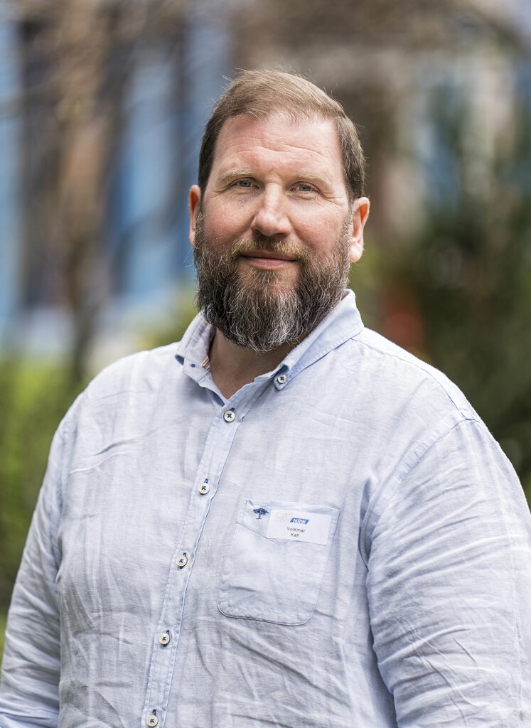 Außenaufnahme eines bärtigen Manns in einem hellblauen Hemd.