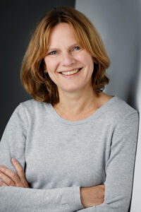 Eine lächelnde Frau mit halblangen Haaren. Sie trägt einen grauen Pullover und hat die Arme verschränkt. 