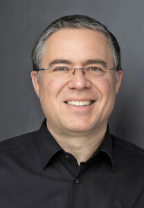 Ein Mann mit Brille in einem schwarzem Hemd vor dunklem Hintergrund