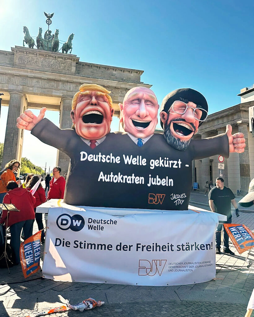 Vor dem Brandenburger Tor und unter strahlend blauem Himmel ist eine Skulptur des Bildhauers Jacques Tilly zu sehen: Trump, Putin und ein Ayatolla als lachende Gestalten, darunter der Schriftzug: "Deutsche Welle gekürzt. Autokraten jubeln."
