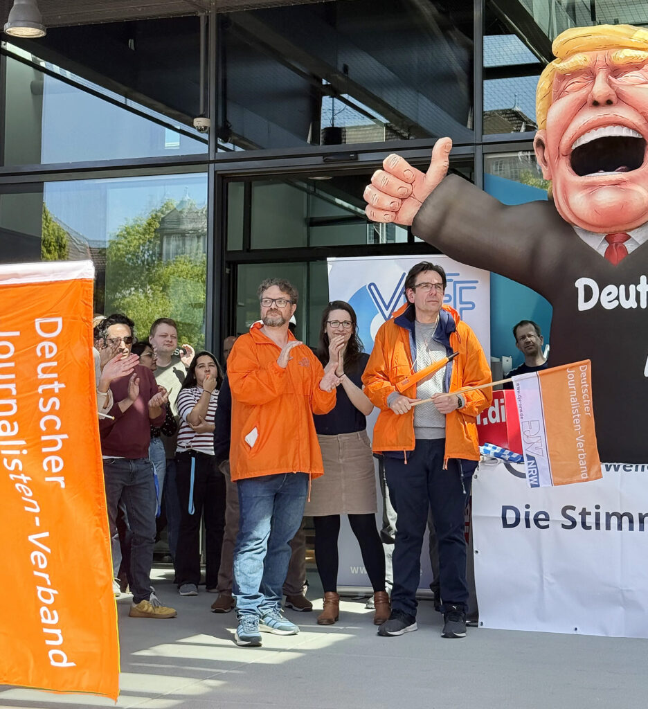 Protest-Veranstaltung bei der Deutschen Welle in Bonn. Zu sehen sind mehreren Menschen, davon mehrere mit organgen Jacken. Daneben Teil der Skulptur des Bildhauers Jacques Tilly mit einer jubelnden Trump-Figur"