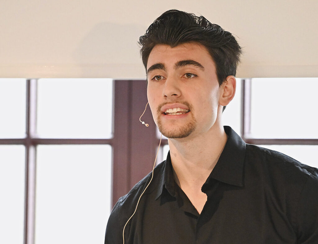 Brustbild eines jungen Mannes mit dunklen Haaren vor einer Fensterfront. Er spricht engagiert in das kleine Mirko eines professionellen Headsets.