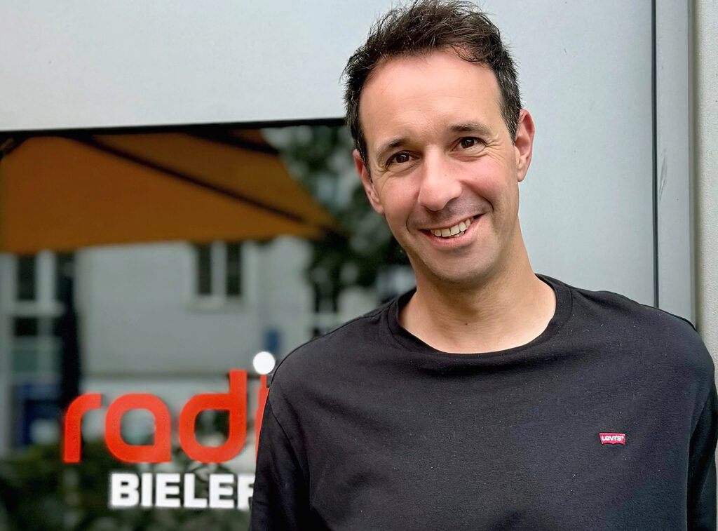 Ein Mann in T-Shirt mit kurzen dunklen Haaren schaut lächelnd in die Kamera. Im Hintergrund ist an einer Fenstrscheibe ein Teil des Schriftzugs "Radio Bildefeld" zu erkennen.
