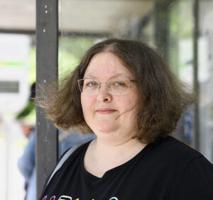 Eine Frau steht im Freien, ihre dunklen kinnlangen Haare wehen leicht im Wind.