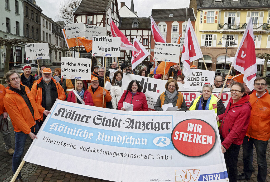 Eine Gruppe von Menschen hat sich mit Fahnen und Transparenten auf einem Platz aufgestellt. Viele von ihnen tragen orange Jacken. Vor ihnen schräg ein großes Banner mit einem dicken Button "Wir streiken". Auf dem Banner stehen die Namen Kölner Stadt-Anzeiger, Kölnische Rundschau, Rheinische Redaktionsgemeinschaft und DJV-NRW.